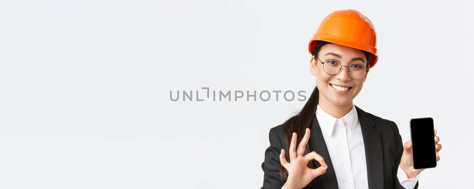 Satisfied young female asian engineer, architect in suit and safety helmet showing mobile phone screen and make okay gesture, approve, recommend application, white background.