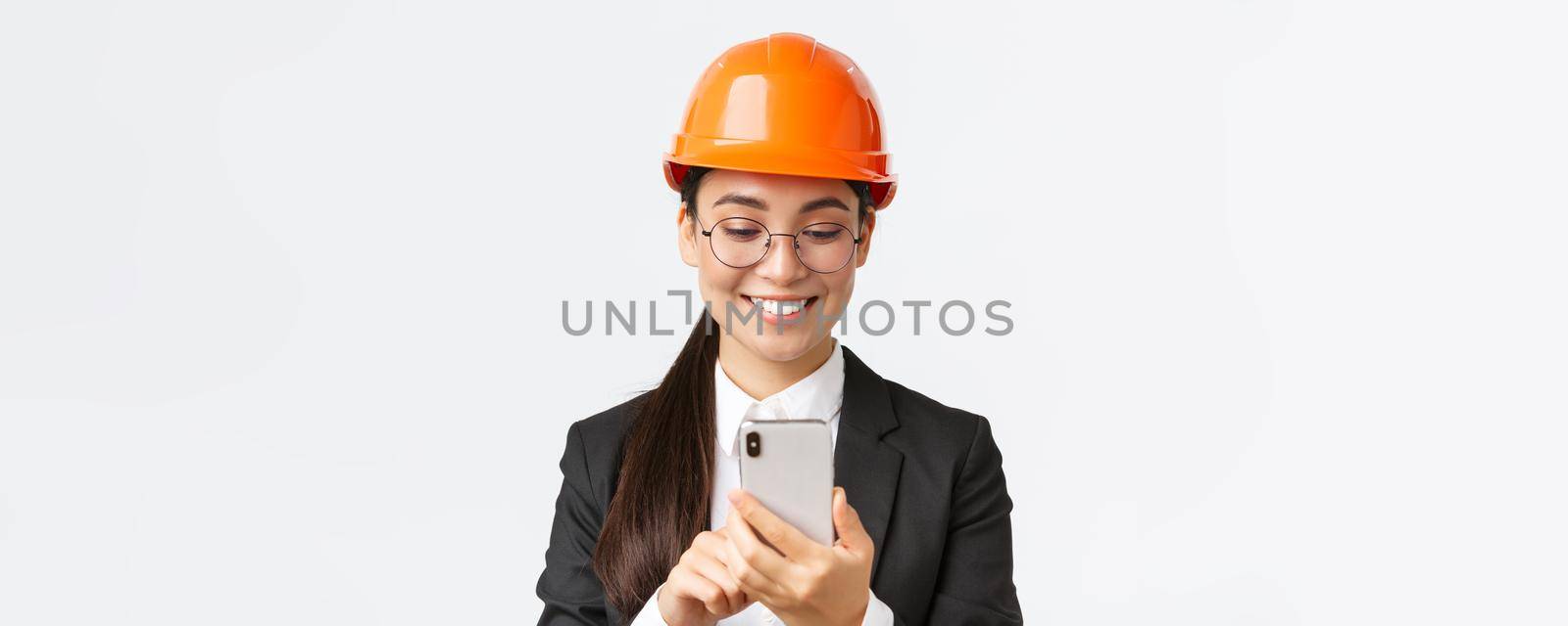Close-up of smiling professional asian female industrial engineer, wearing safety helmet and business suit, using smartphone, looking pleased at mobile phone screen, standing white background by Benzoix