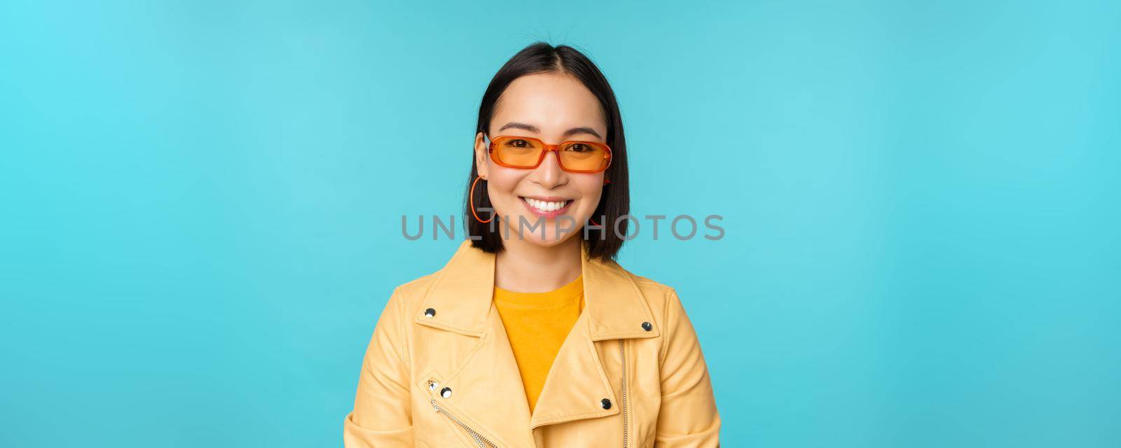 Close up portrait of smiling stylish asian woman in sunglasses, looking happy at camera, posing in trendy yellow jacket, blue background.