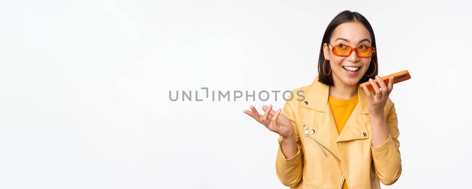 Image of happy asian girl talking on speakerphone, recording, translating her voice with mobile phone app, talking in smartphone dynamic, standing over white background.