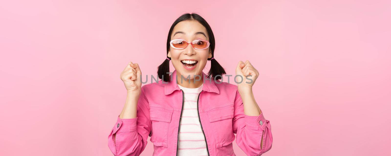 Portrait of excited japanese girl in sunglasses, celebrating, achieve goal, gasping amazed and smiling, standing over pink background.