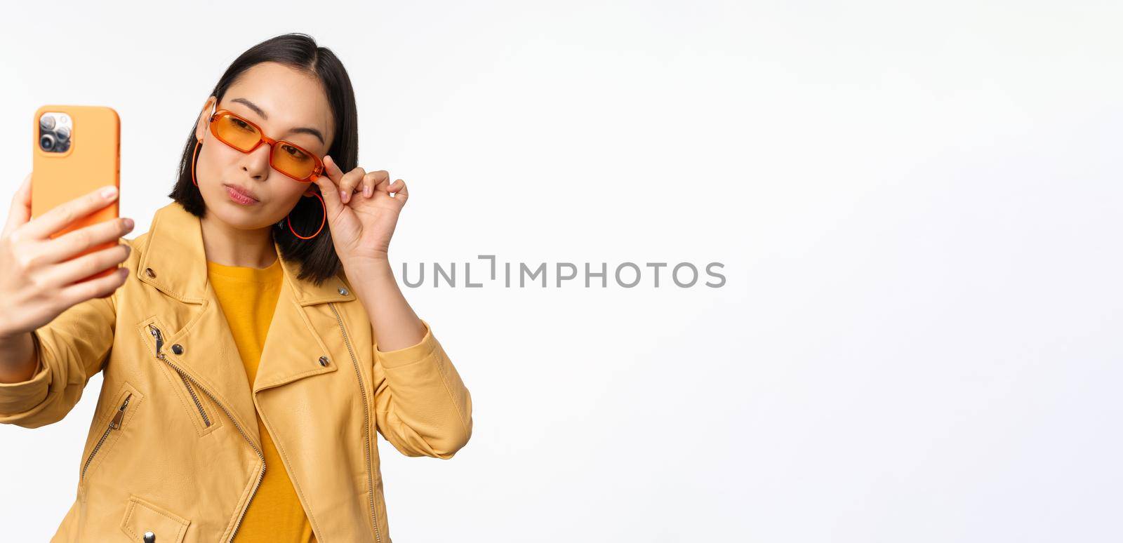 Stylish, beautiful asian girl in sunglasses, taking selfie on smartphone, posing for photo, holding mobile phone, white studio background.