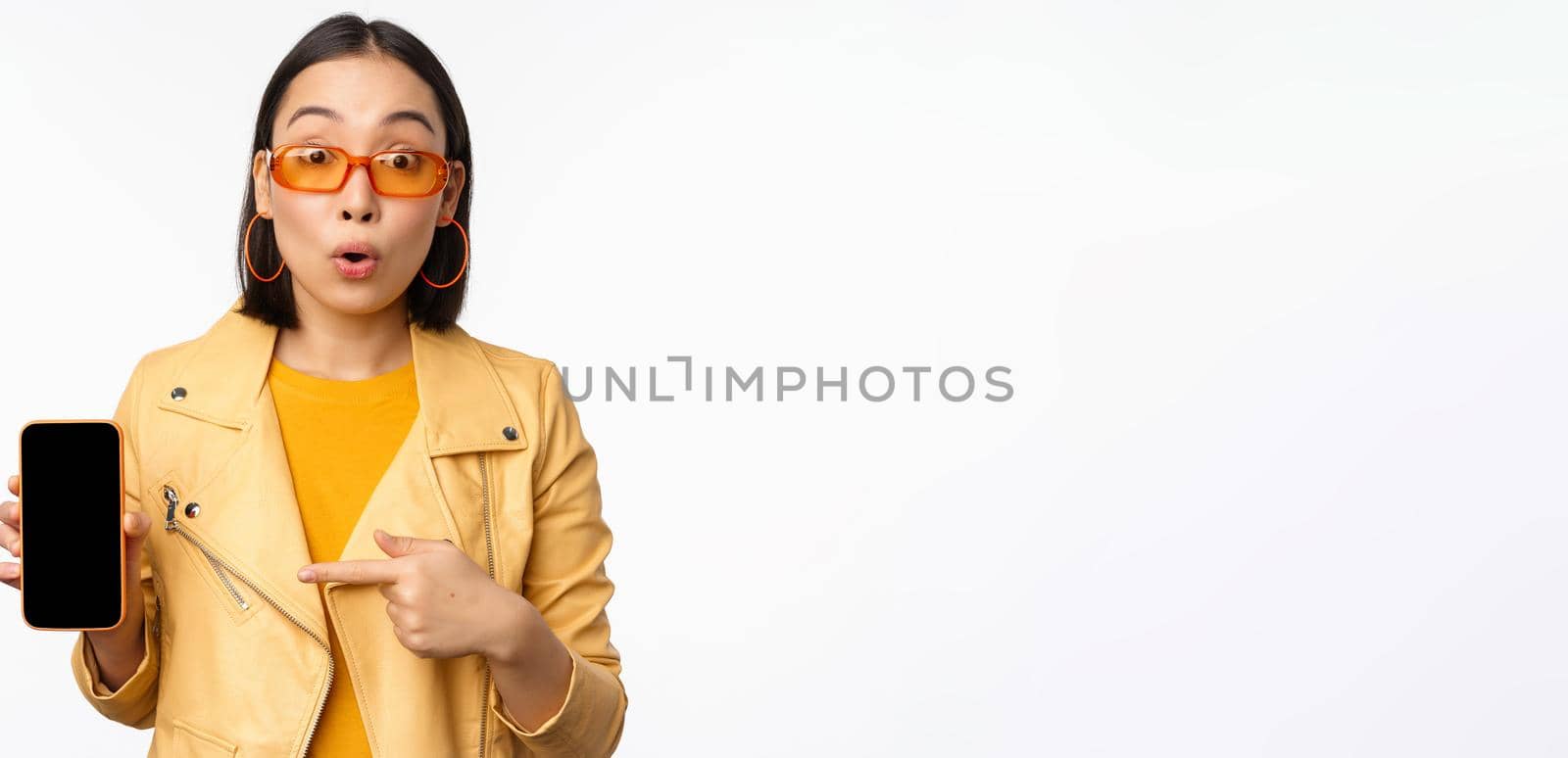 Portrait of stylish korean girl in sunglasses, smiling, pointing finger at smartphone screen, showing mobile phone application, standing over white background.