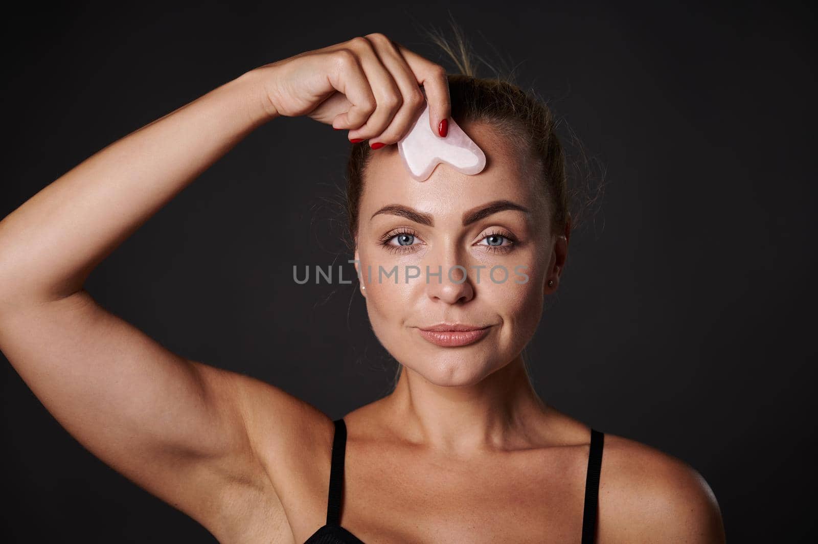 Studio shot. Beauty portrait of an attractive Caucasian woman massaging her face with a gua-sha jade stone massager. Anti-aging, anti-wrinkles, rejuvenating and anti-puffiness concept