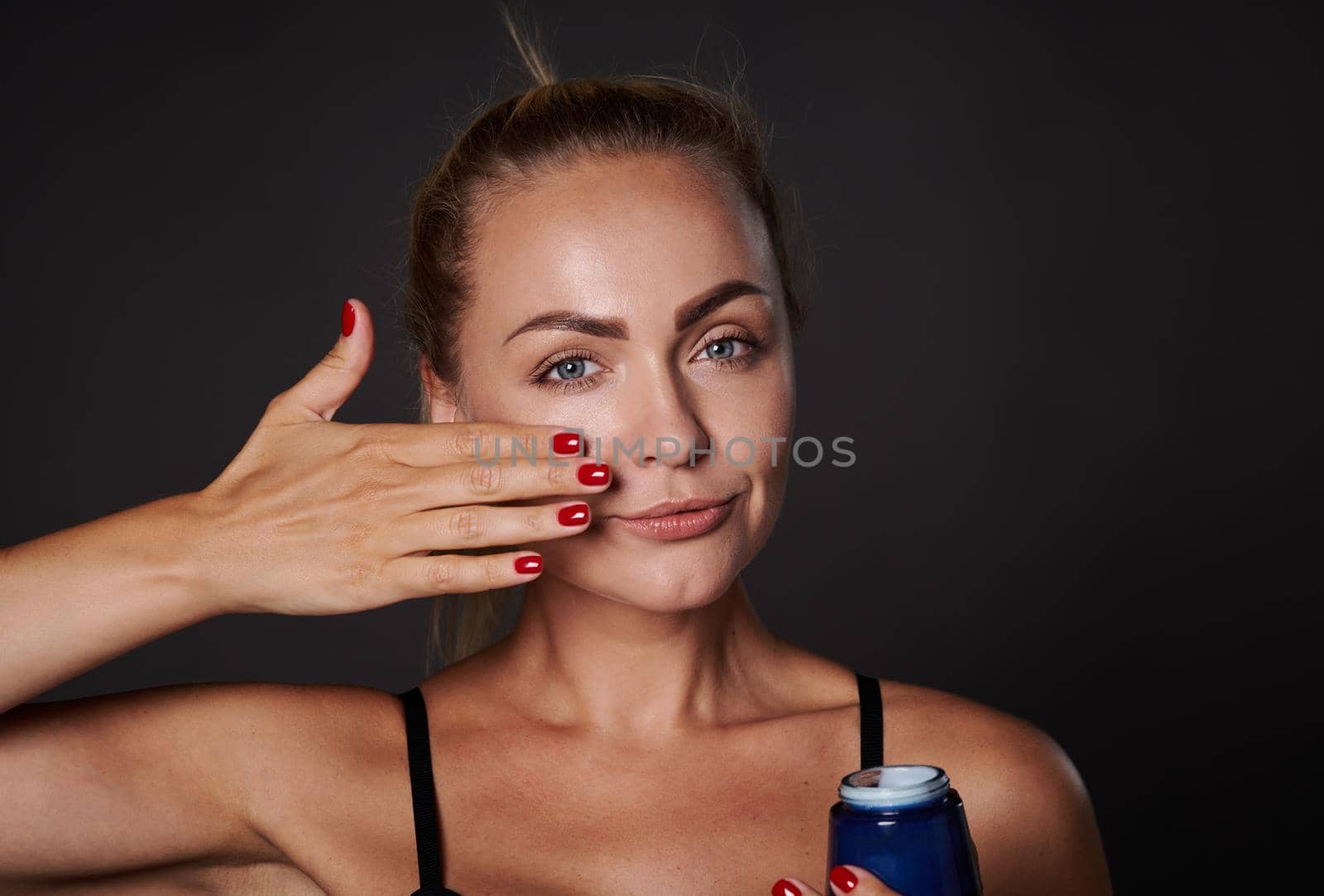 Close-up beauty portrait beautiful Caucasian woman applying moisturizer on face, holding jar of daily smoothing cream or beauty serum, isolated on black background. Skin care, cosmetology, spa concept by artgf