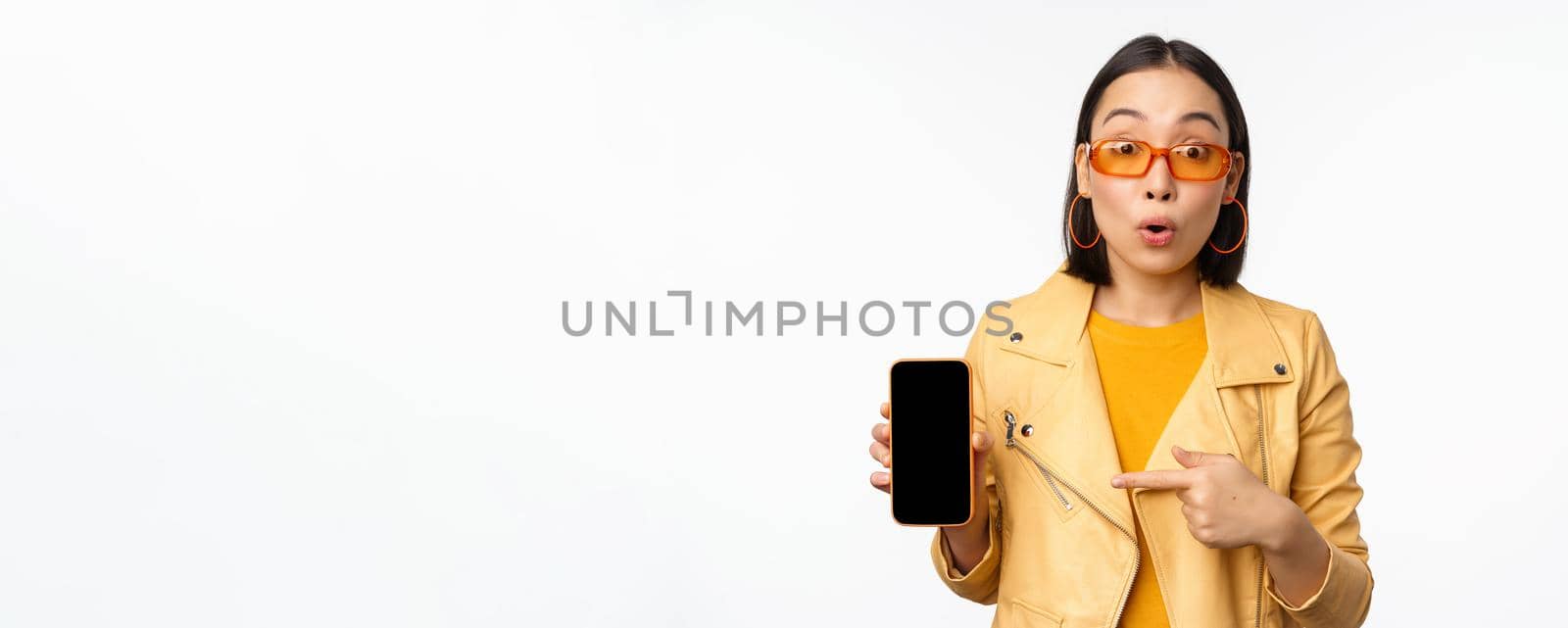 Portrait of stylish korean girl in sunglasses, smiling, pointing finger at smartphone screen, showing mobile phone application, standing over white background.