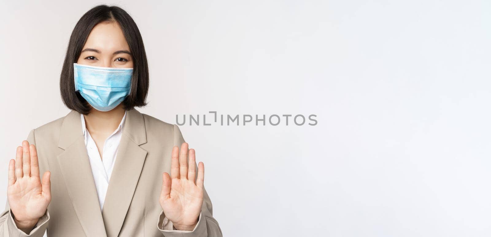 Coronavirus and work concept. Portrait of asian female office lady, woman at workplace wearing medical face mask and showing stop, prohibition gesture, white background.