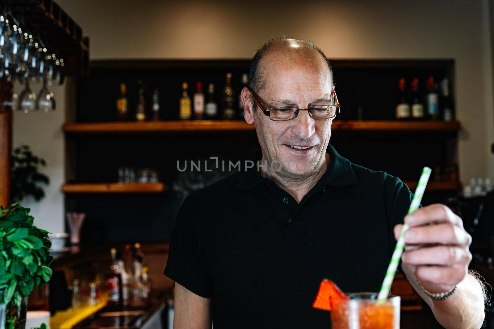 experienced waiter, concentrated and precise, dressed in company uniform, black polo shirt, stirring a cocktail in a crystal glass on the counter of the nightclub. Preparing cocktails for customers. Warm atmosphere and dim light. Horizontal