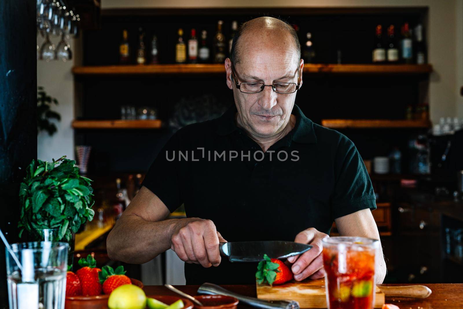 bald adult male, experienced, concentrated and hard-working waiter, dressed in company uniform, black polo shirt, cutting strawberries for a cocktail at the nightclub counter. Preparing cocktails for the customers. Warm atmosphere and dim light. Horizontal