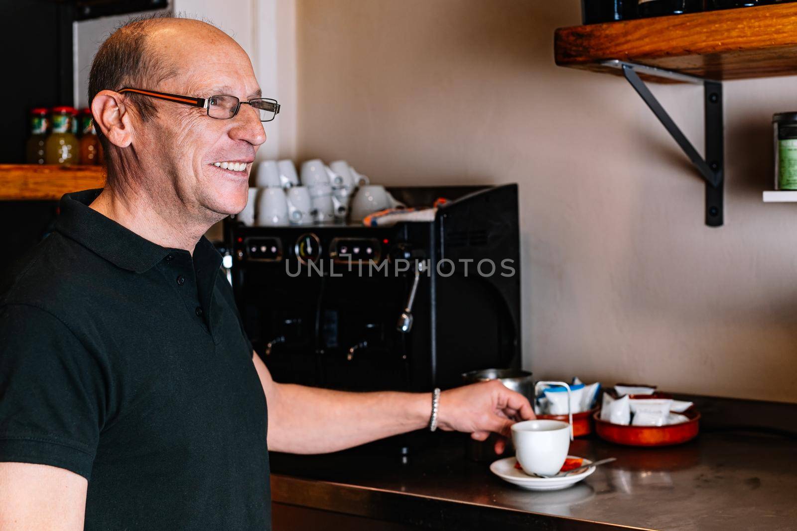 Male waiter, mature, bald, smiling, dressed in company uniform, black polo shirt, Mature waiter preparing a coffee in the coffee machine. Waiter talking to his co-worker. Warm atmosphere and dim lighting. Horizontal