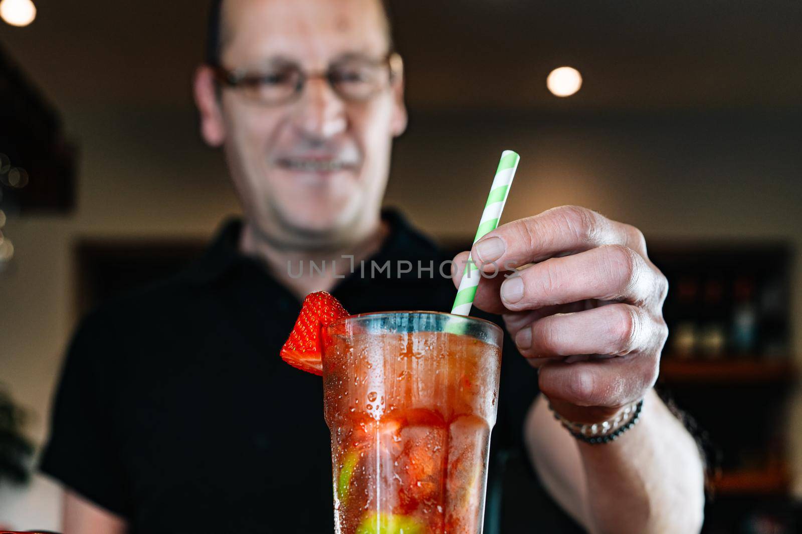 detail of the hands of an experienced waiter, concentrated and precise, dressed in company uniform, black polo shirt, stirring a cocktail in a crystal glass at the nightclub counter. Preparing cocktails for the customers. Warm atmosphere and dim light. Horizontal. Low angle view,