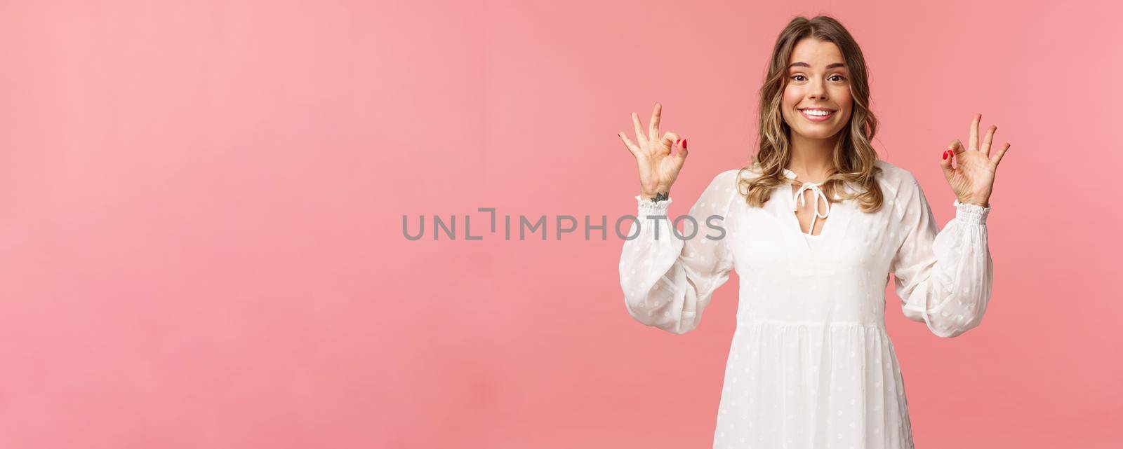Beauty, fashion and women concept. Portrait of excited and upbeat blond female in white dress, standing satisfied over pink background, show okay sign, guarantee and assure in quality.