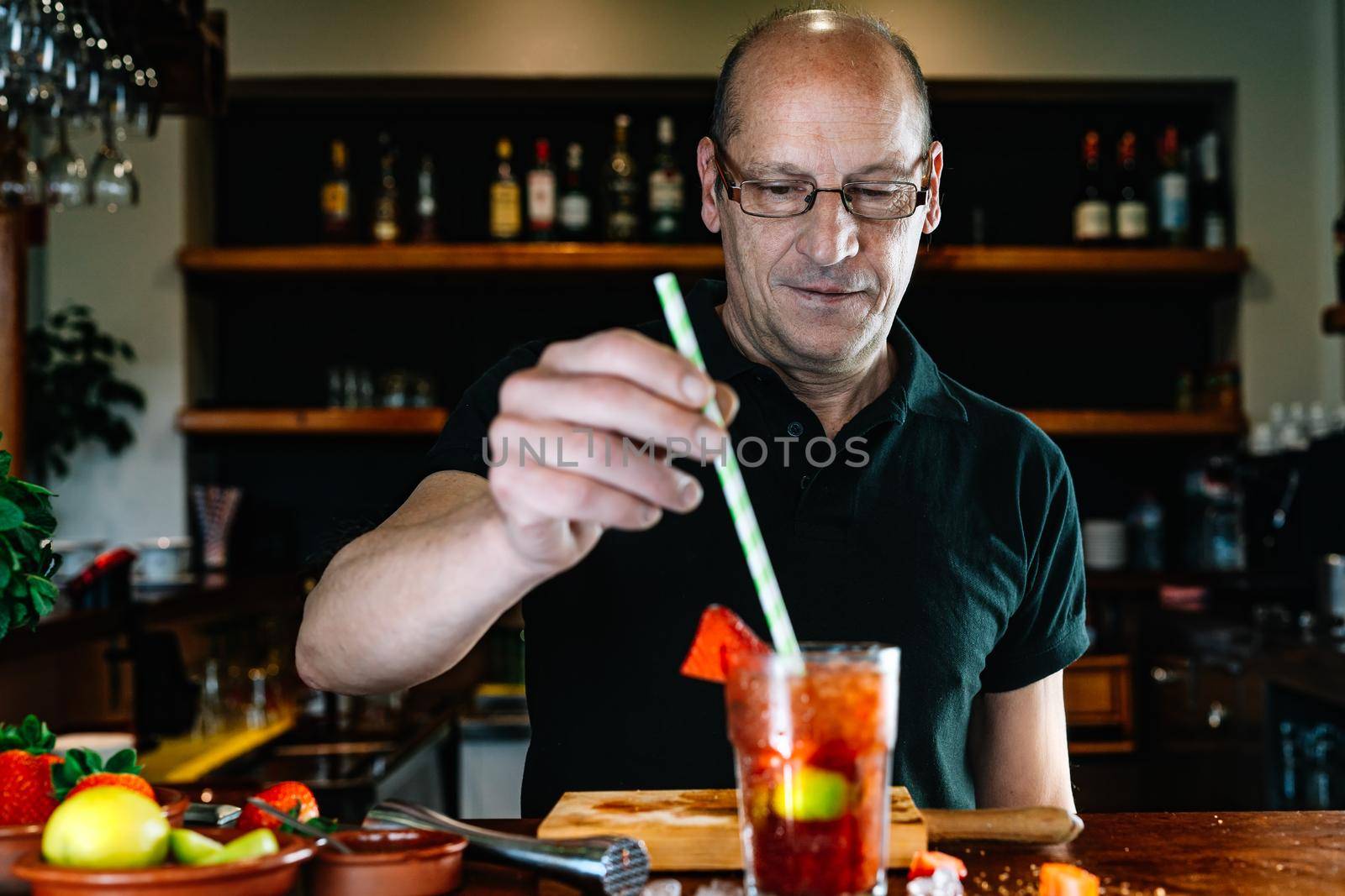 experienced waiter, concentrated and precise, dressed in company uniform, black polo shirt, stirring a cocktail in a crystal glass on the counter of the nightclub. Preparing cocktails for customers. Warm atmosphere and dim light. Horizontal