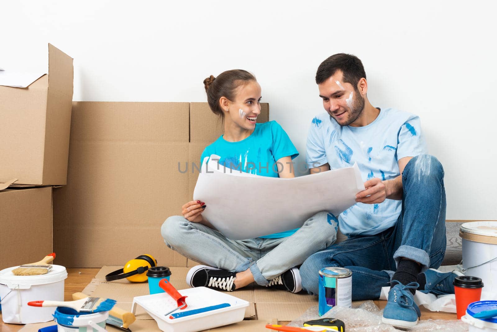 Couple discussing renovating plan of their new home. Smiling young man and woman together sitting on floor among cardboard boxes. House remodeling and interior redesign. People looking at blueprint.