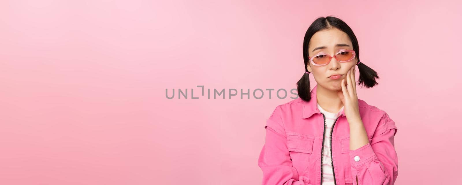 Image of sad asian girl sulking, touching her cheek, pouting disappointed, has toothache, standing over pink background.