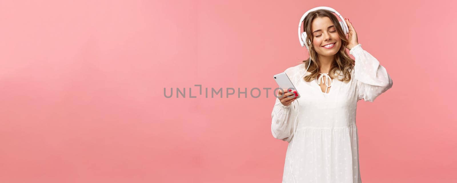 Portrait of tender happy and cute blond girl in white dress, close eyes and smiling pleased as listening music in headphones, feeling warmth and happiness enjoying favorite track, hold mobile phone.