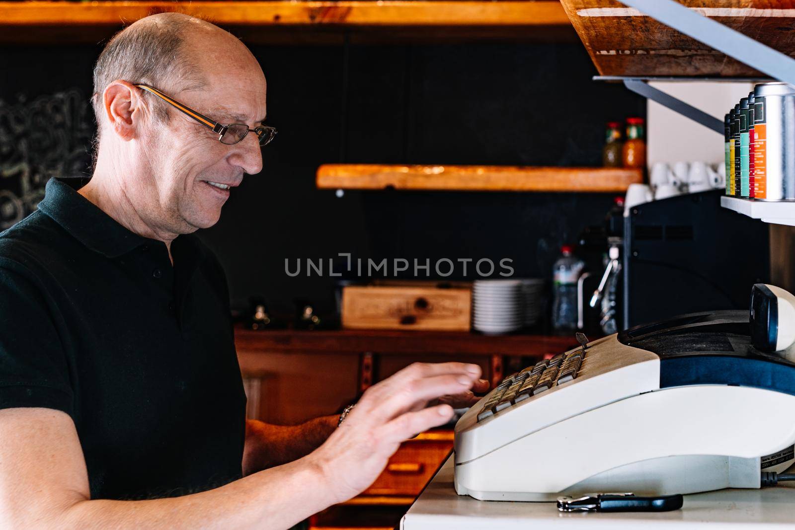 Adult male waiter, bald with glasses, smiling, experienced, focused and hardworking, dressed in company uniform, a black polo shirt, collecting a service at the cash register. Waiter registering an order. Warm atmosphere and dim lighting. Horizontal