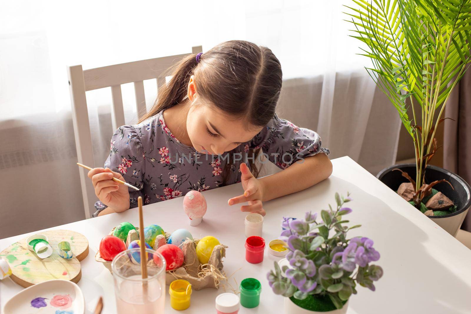 A cute little girl sits at a white table near the window, prepares for the Easter holiday, paints eggs with a brush.