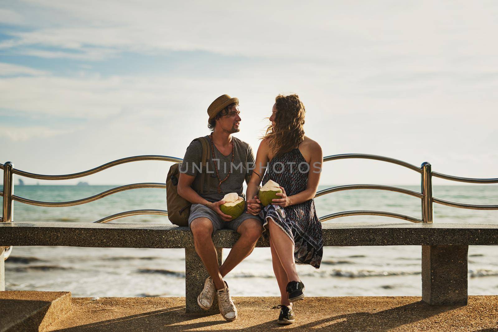 Chatting by the seaside. Shot of a happy young couple enjoying cocktails while relaxing on a bench by the beach. by YuriArcurs