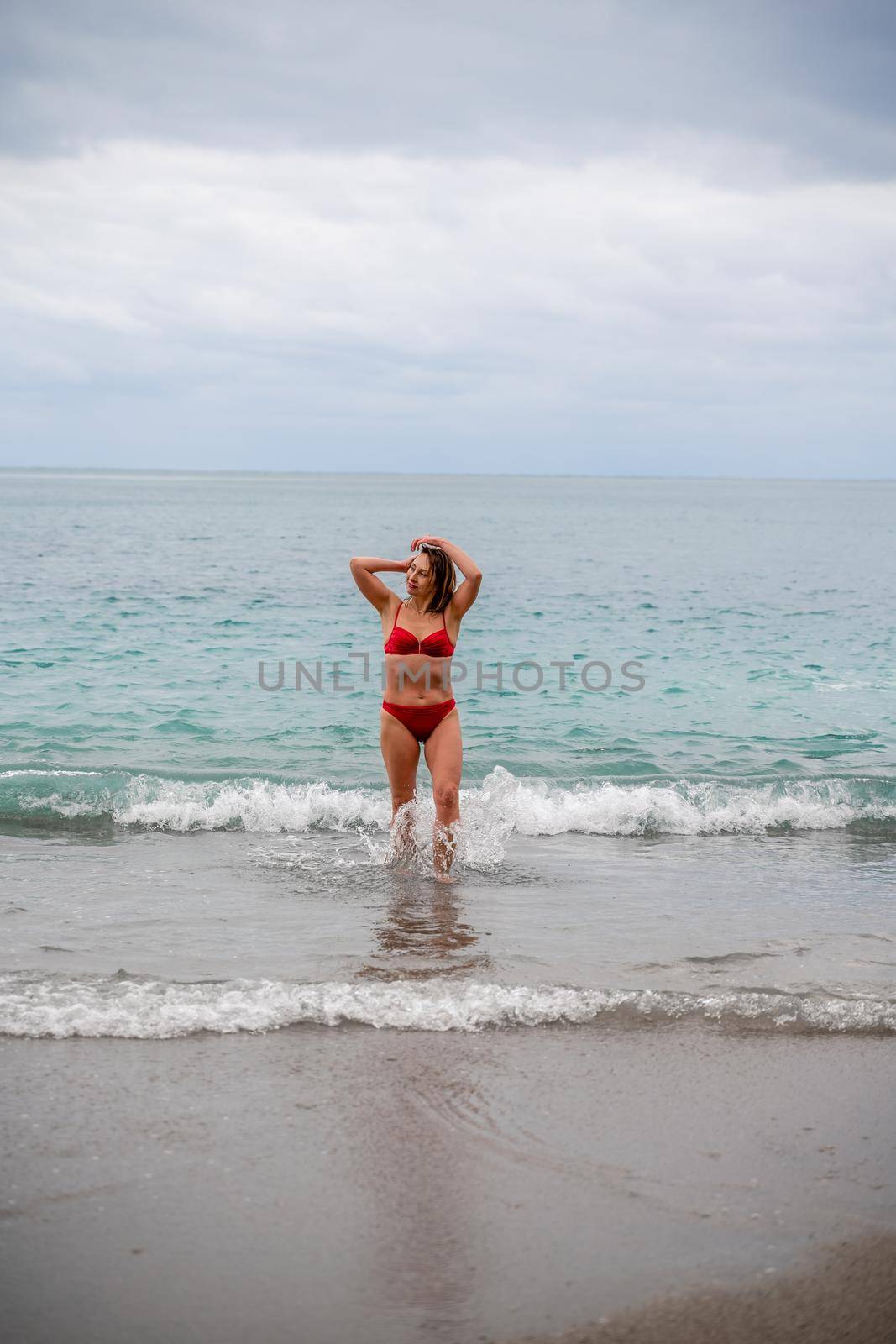 A middle-aged woman with a good figure in a red swimsuit on a pebble beach, running along the shore in the foam of the waves.