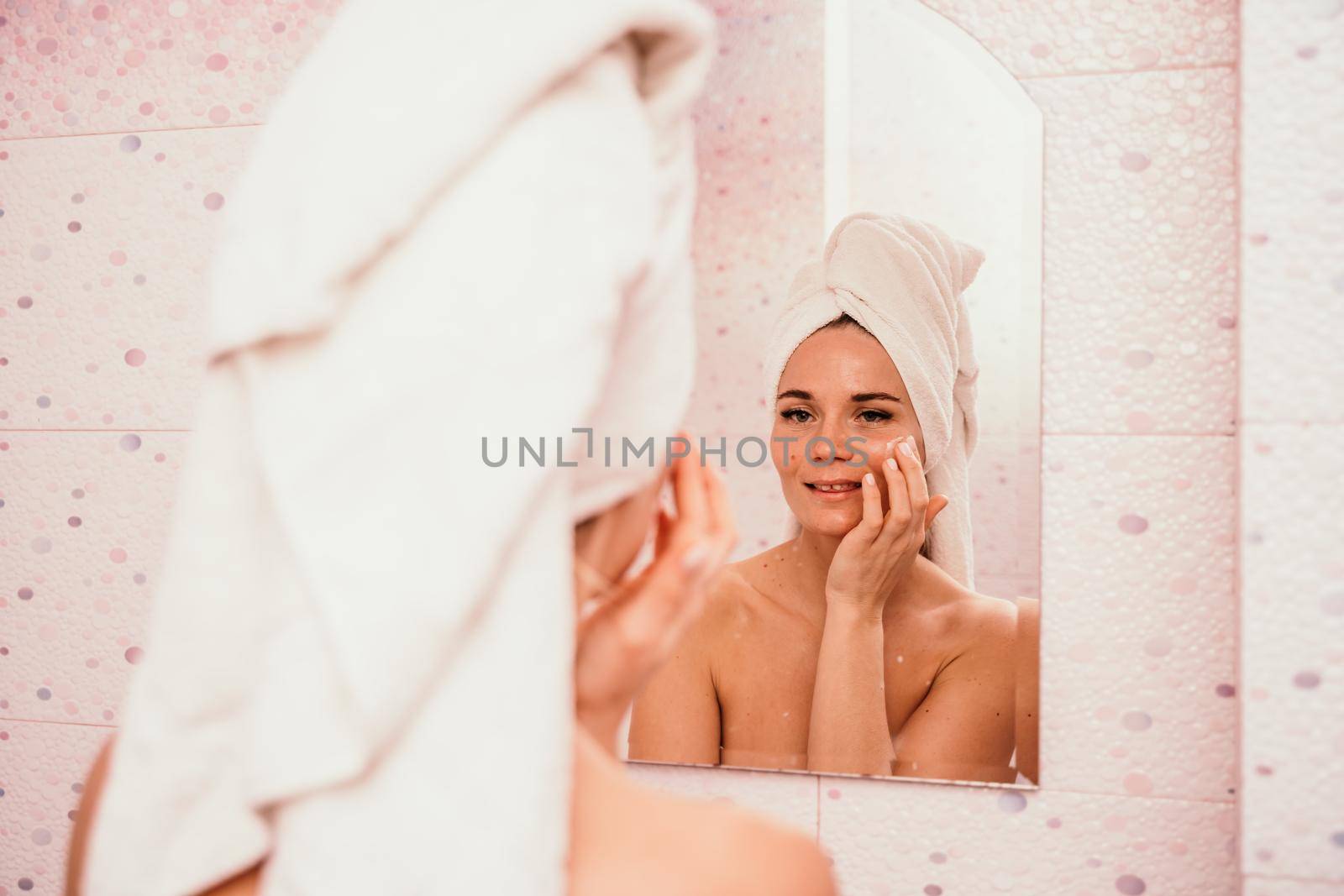 Young beautiful woman using face cream moisturizing lotion after bath. Pretty attractive girl in a towel on her head stands in front of a mirror in a home bathroom. Daily hygiene and skin care.