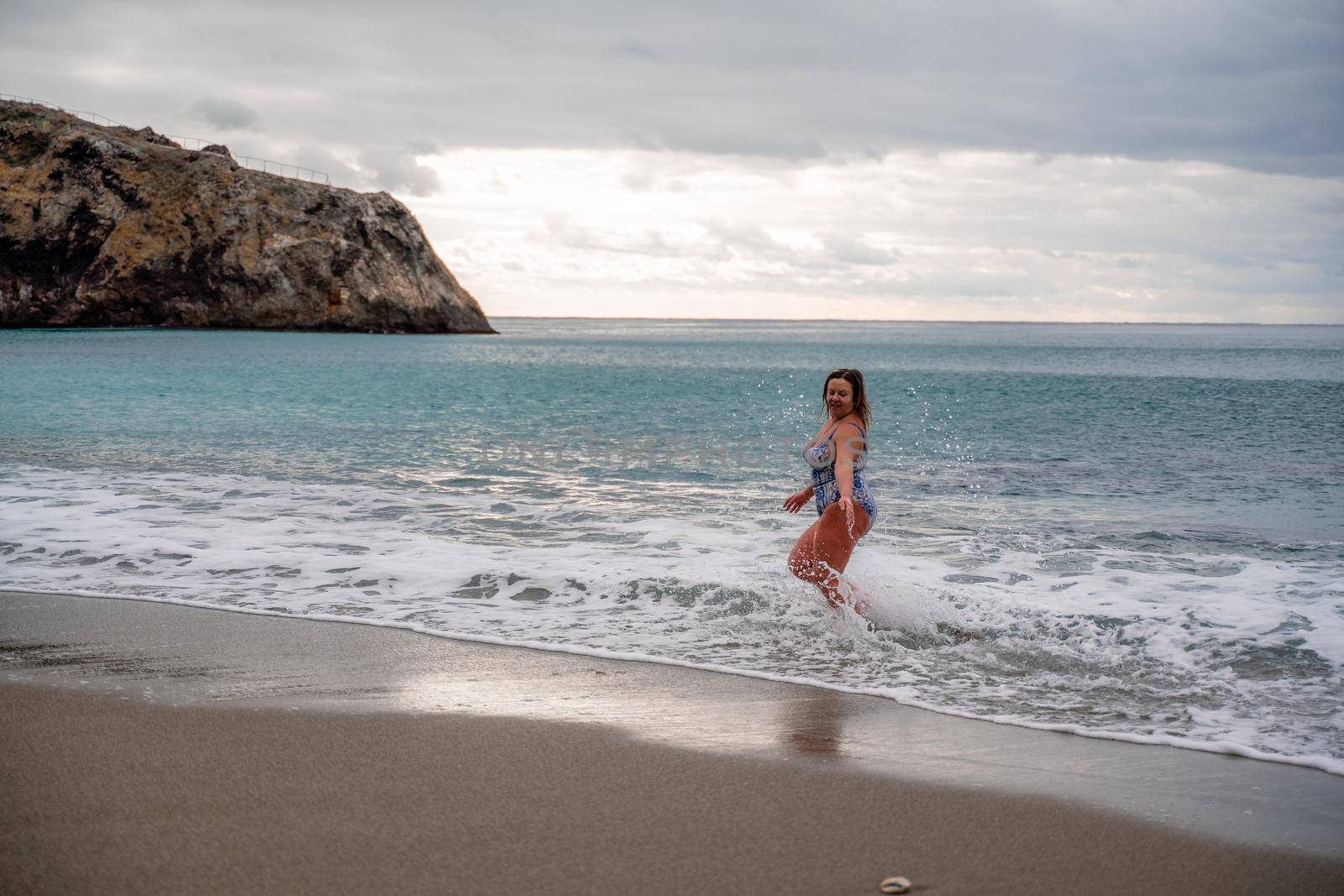 A plump woman in a bathing suit enters the water during the surf. Alone on the beach, Gray sky in the clouds, swimming in winter