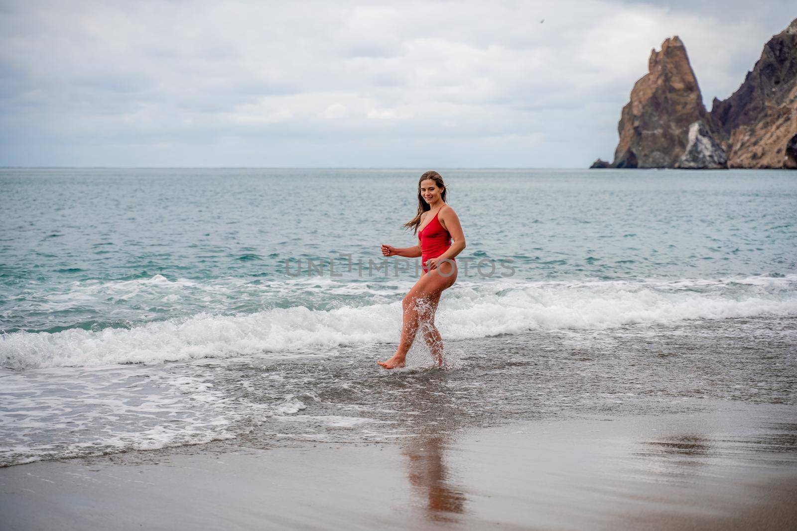 A beautiful and sexy brunette in a red swimsuit on a pebble beach, Running along the shore in the foam of the waves by Matiunina