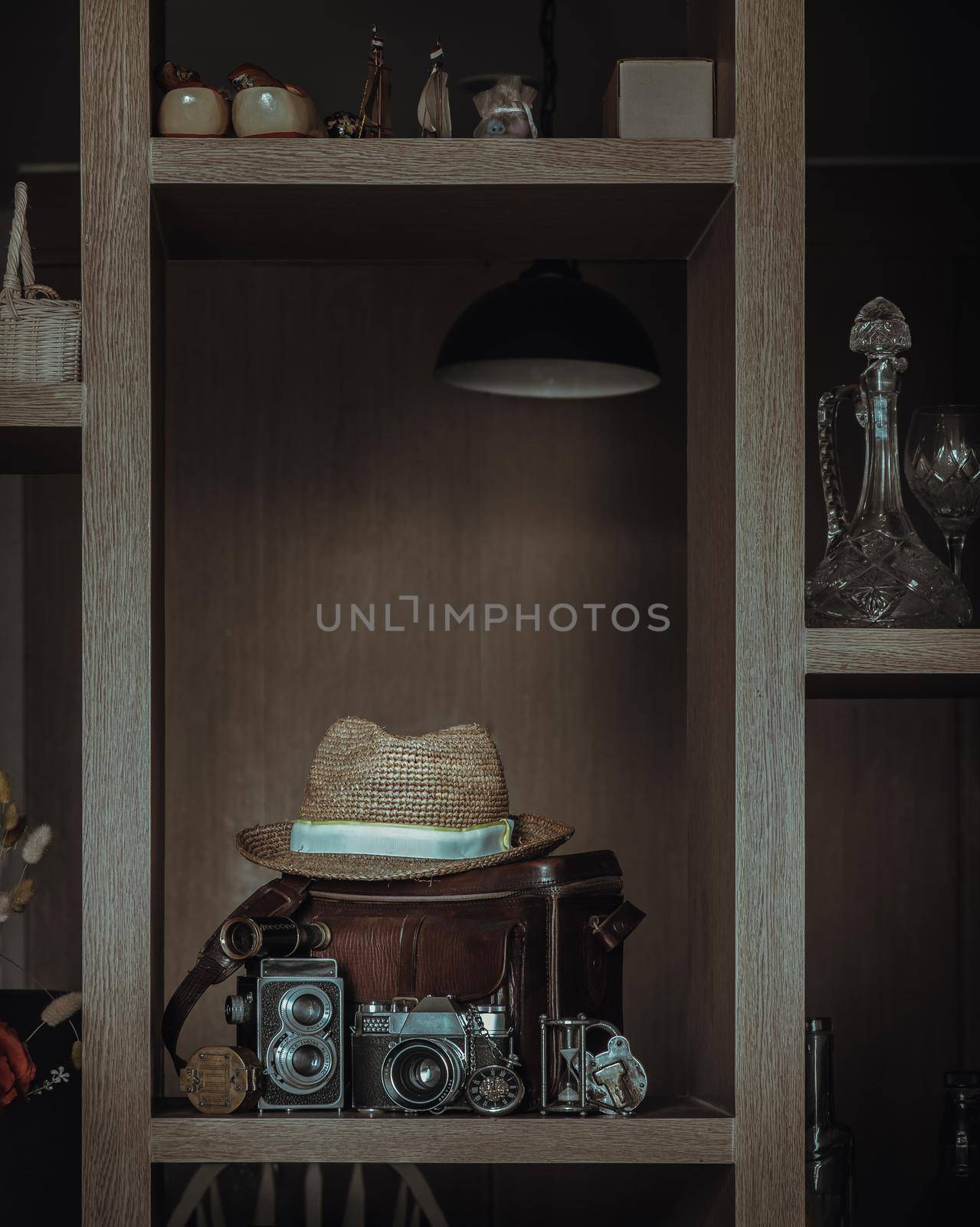 Wooden shelves displaying vintage style collectibles have Hourglass, Retro pocket watch, Binocular, Compass, iron padlock, Photo camera and Straw fedora hat on Leather bag in the living room. Interior style concept. No focus, specifically.