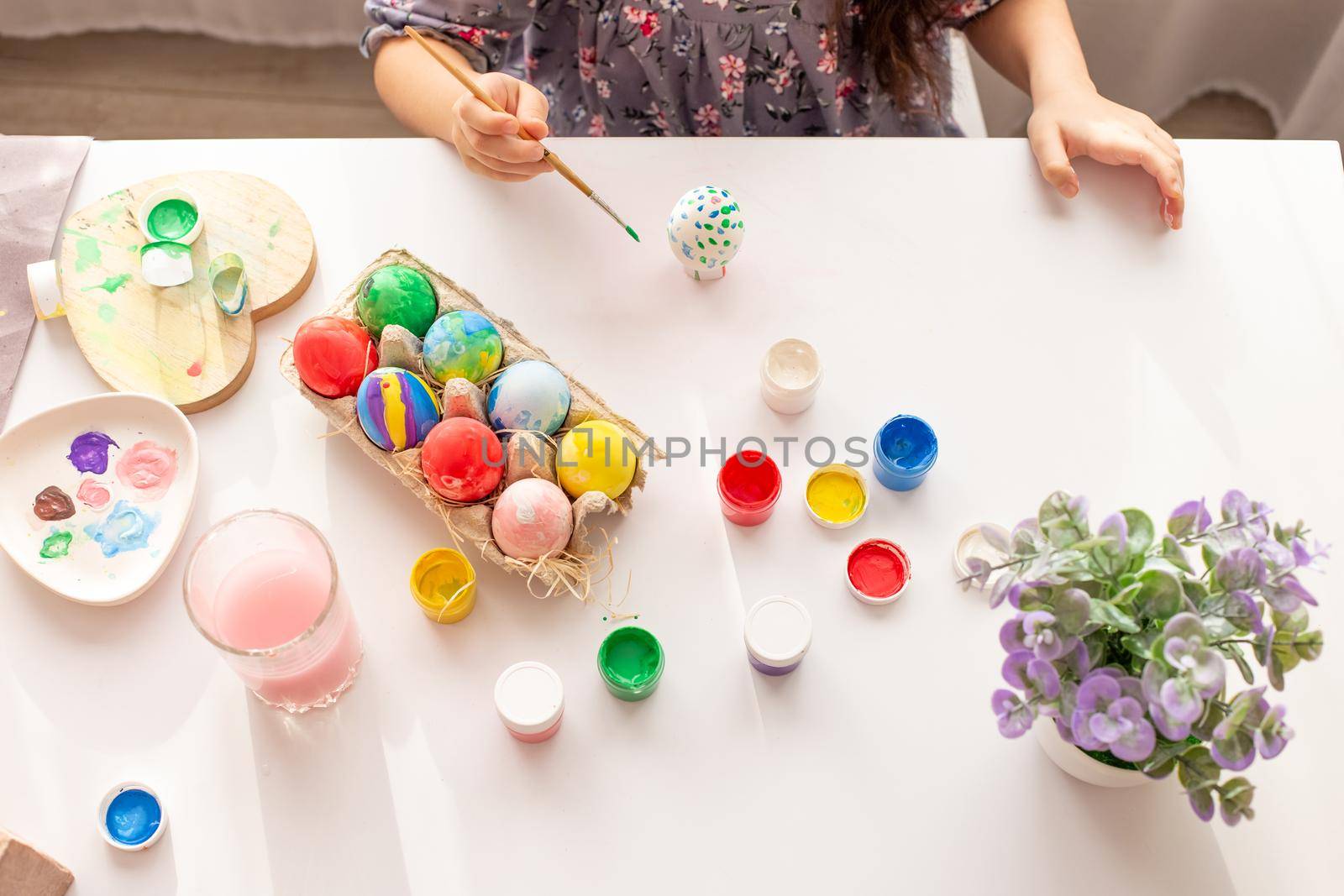 Hands of a little girl, with a painted egg, on a white table with multi-colored eggs in a tray, brushes and paints. Top view