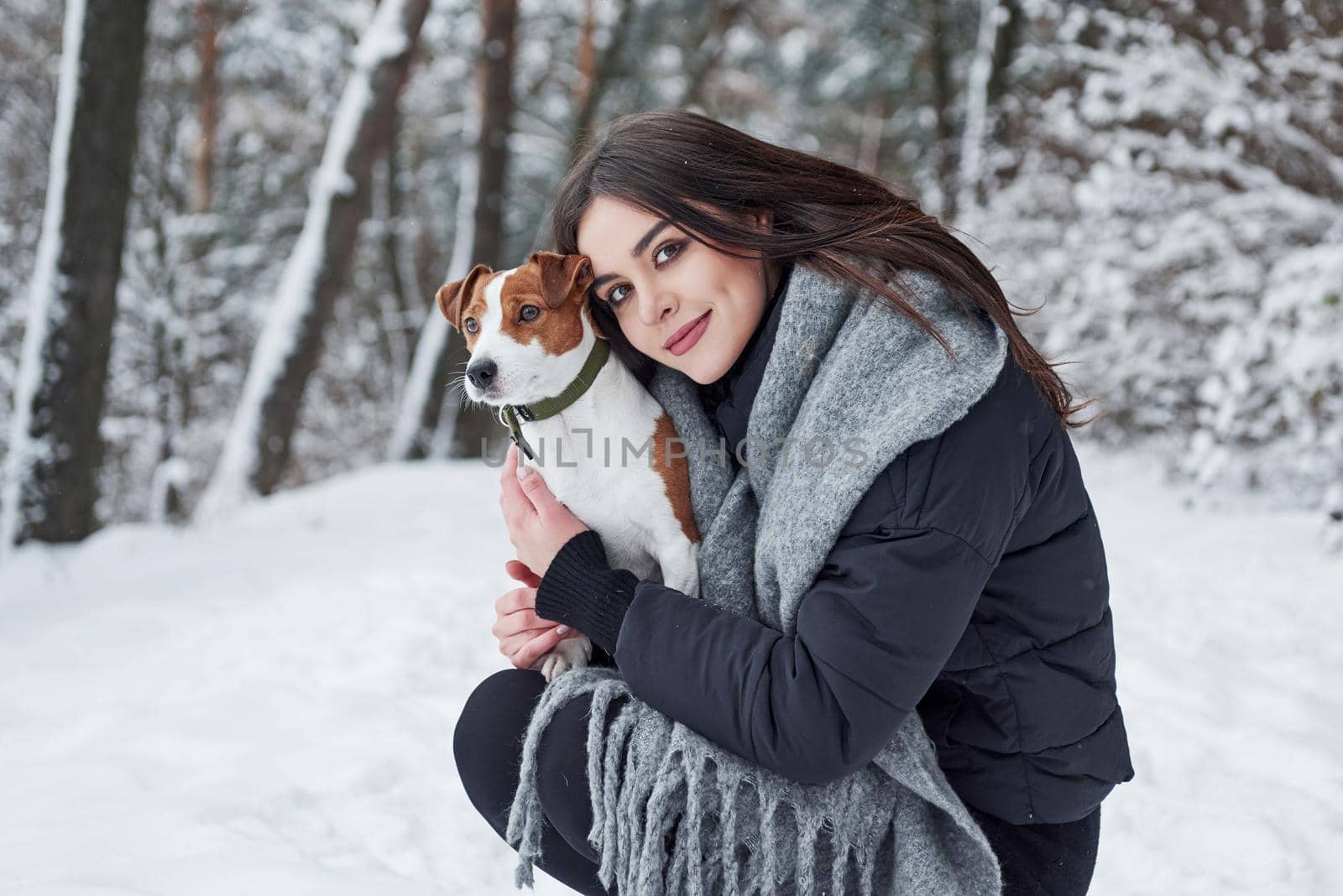 Girl looking to the camera, pet to the side. Smiling brunette having fun while walking with her dog in the winter park.