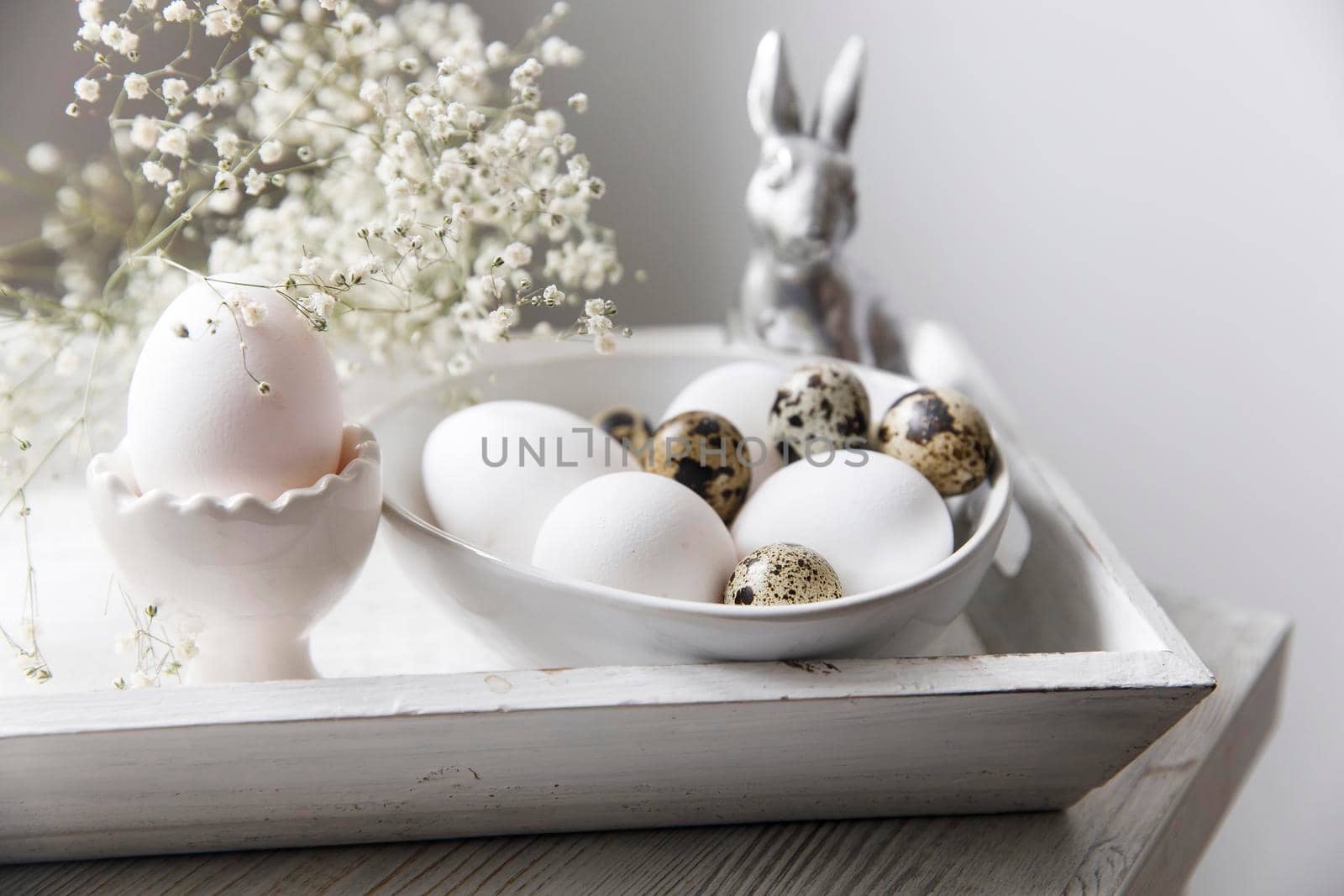 Table decorated for Easter in beige tones. Dried gypsophila, chicken and quail eggs, a silver figurine of a hare and a cup of coffee. Easter concept. by elenarostunova