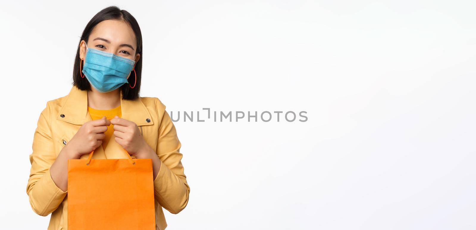 Covid-19 and shop concept. Young asian stylish woman, wearing medical face mask, holding shopping bag, going in malls during pandemic, white background.