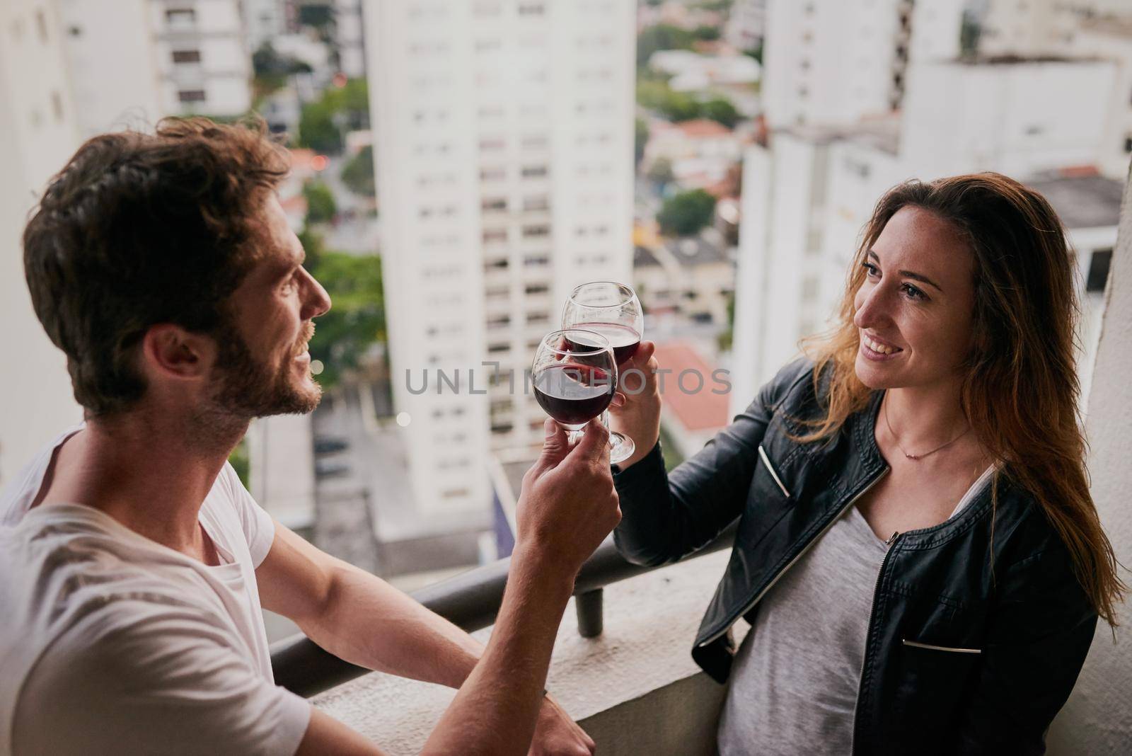 Celebrating our adventure through the city. Cropped shot of an attractive young couple drinking wine on a rooftop looking over the city. by YuriArcurs