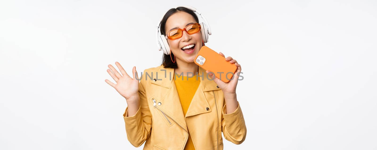 Happy stylish asian woman in sunglasses, listening music in headphones, holding smartphone, singing and dancing, standing over white background.