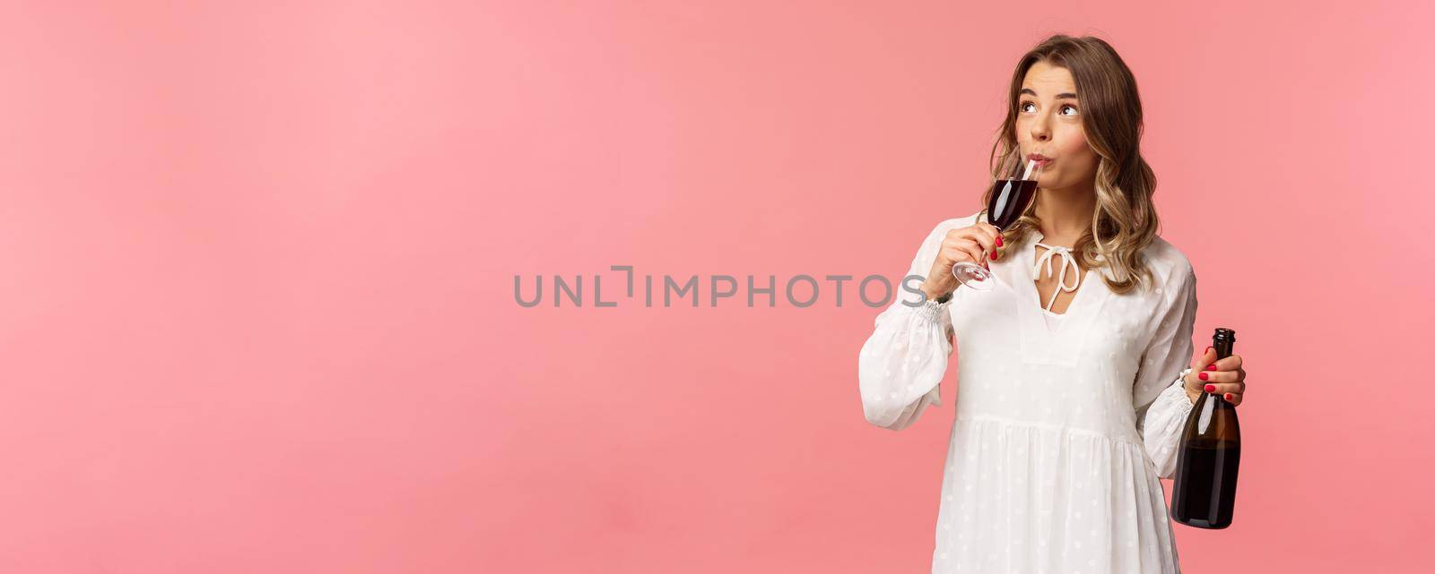 Holidays, spring and party concept. Portrait of carefree independent cute blond woman sipping wine from glass, holding bottle and tasting drink, look up, celebrating with friends, pink background by Benzoix