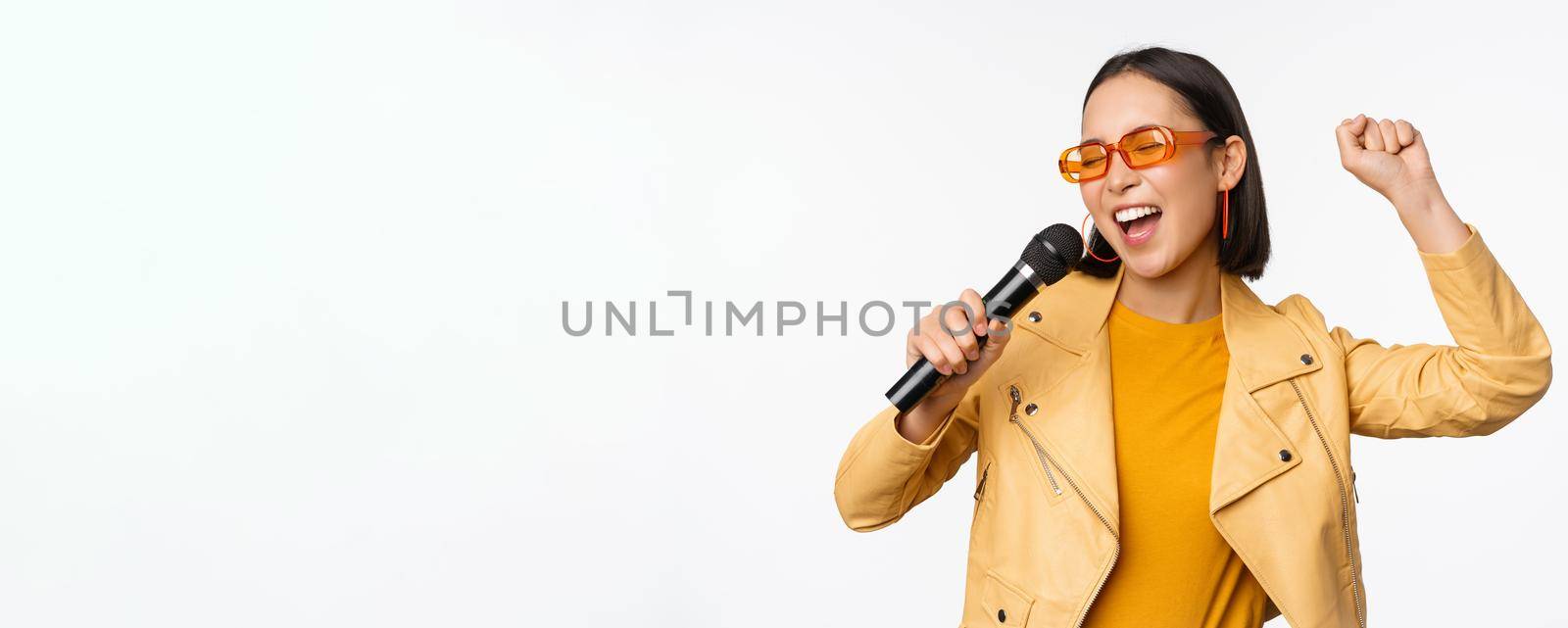 Stylish asian girl in sunglasses, singing songs with microphone, holding mic and dancing at karaoke, posing against white background.
