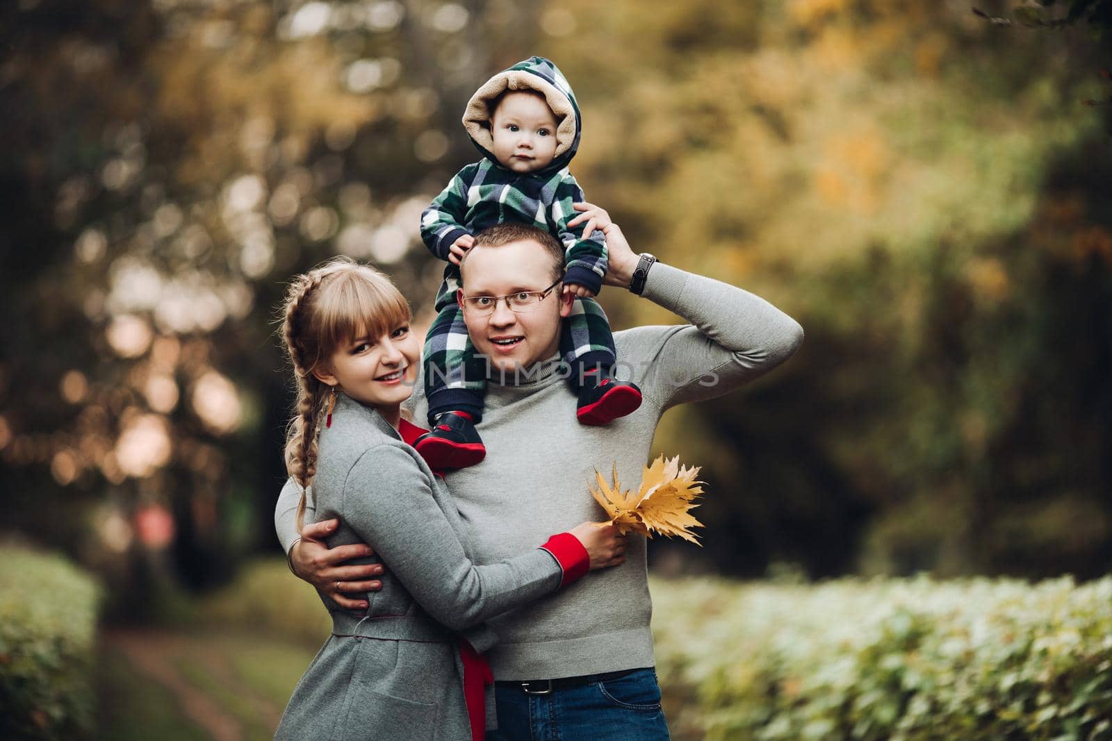 Portrait of attractive young mother and handsome smiling father wearing glasses holding their beautiful lovely baby girl on hands standing against green hedge in autumnal park. They are smiling and looking at camera.