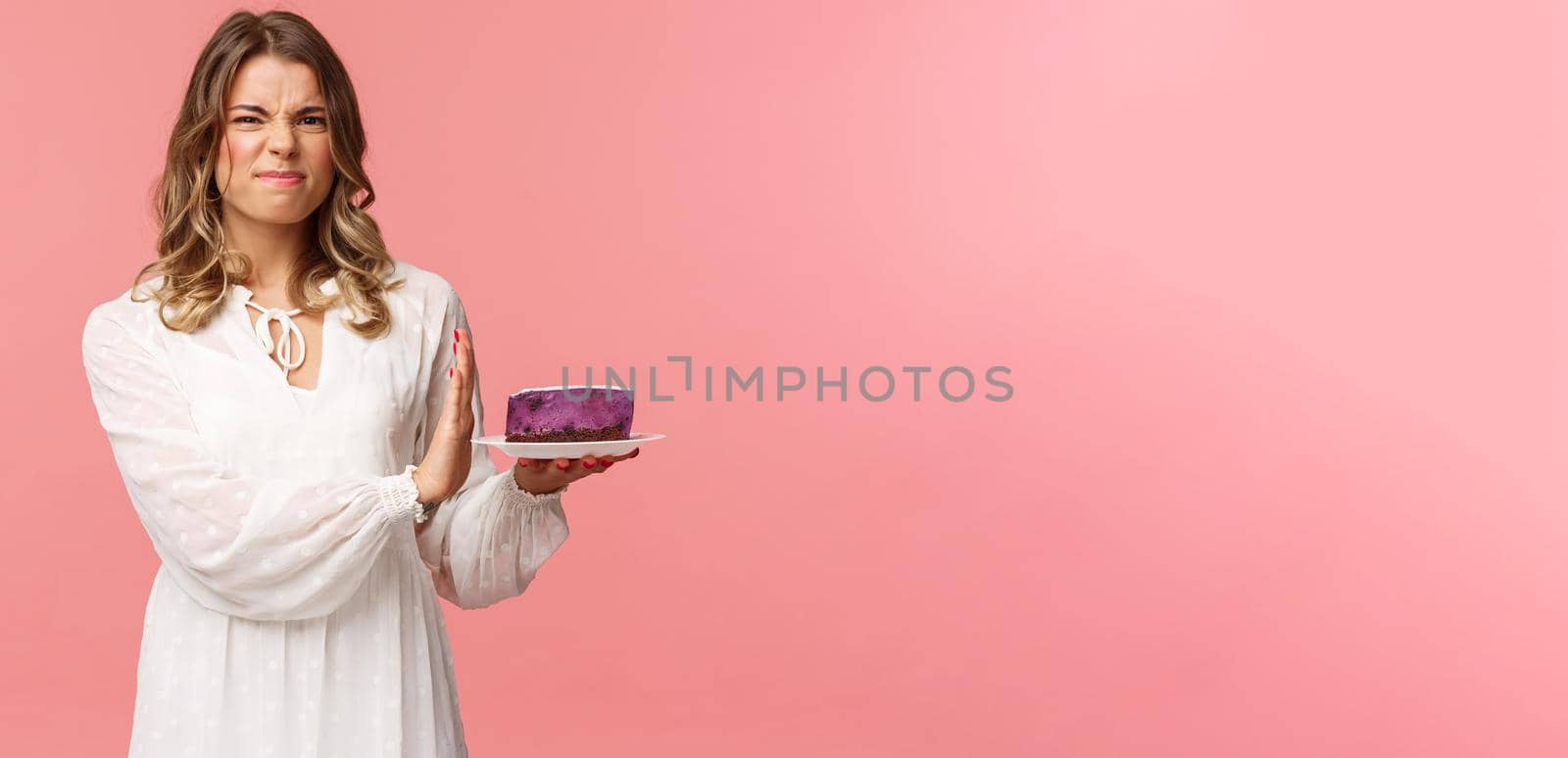Holidays, spring and party concept. Portrait of dissatisfied and displeased blond girl in white dress rejecting, declining eat tasteless cake, grimacing and show stop sign at dessert, pink background.