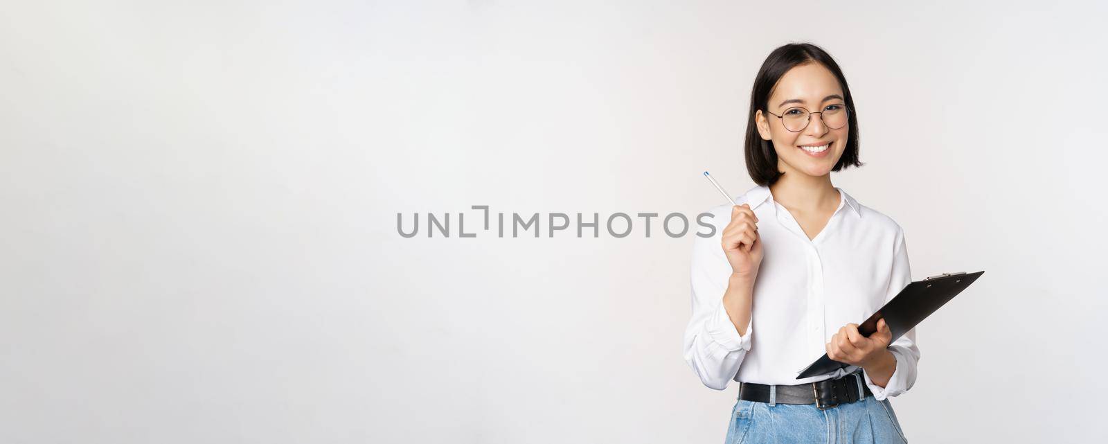 Smiling young asian woman taking notes with pen on clipboard, looking happy, standing against white background.