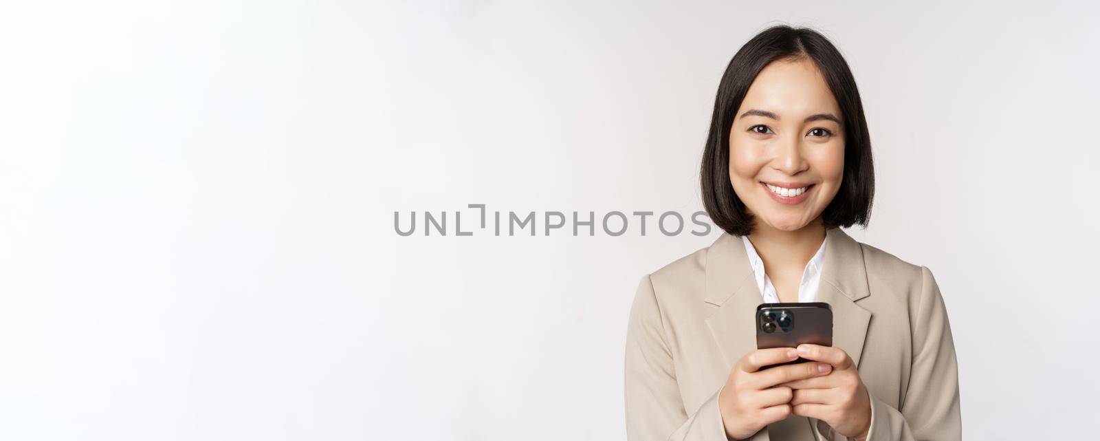 Image of asian businesswoman in suit, holding mobile phone, using smartphone app, smiling at camera, white background.