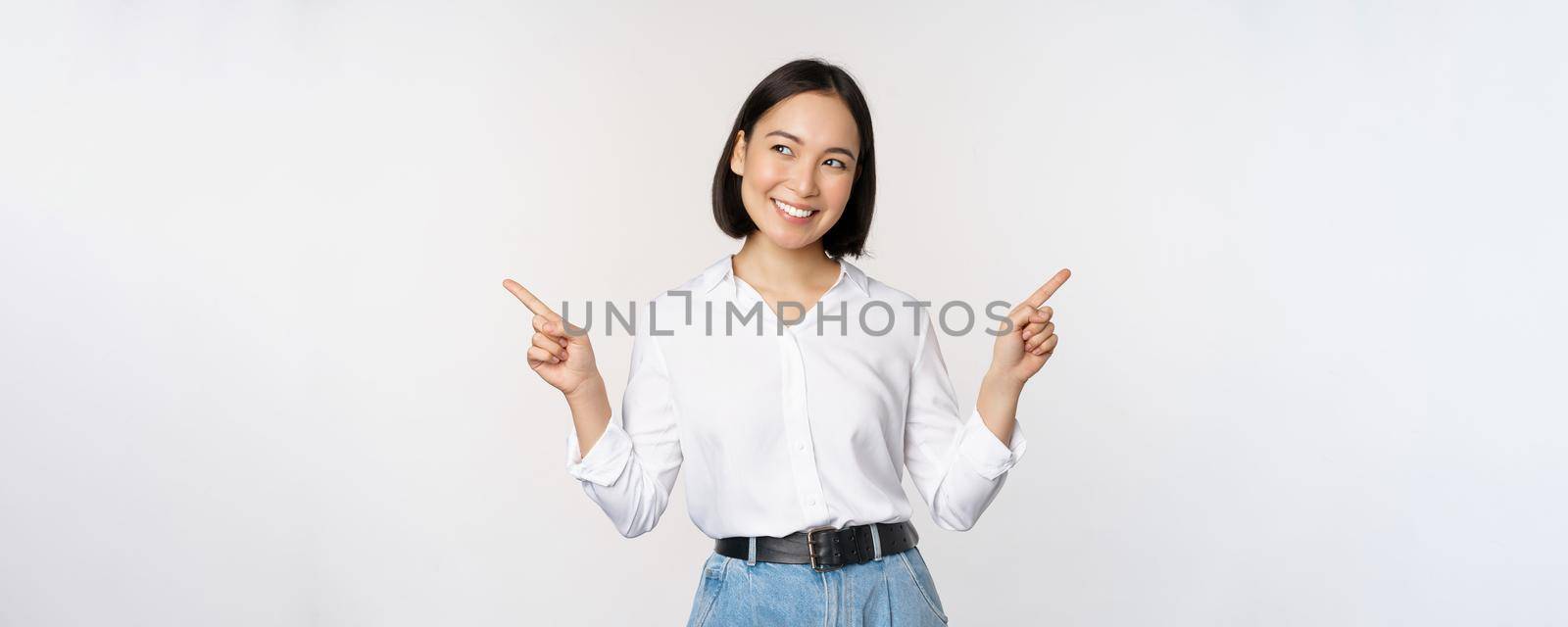 Image of beautiful asian woman pointing fingers left and right, making decision, showing two variants choices, standing over white background by Benzoix
