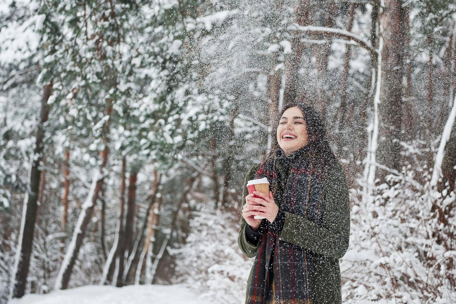 Smiles when snow falling from the trees on her. Girl in warm clothes with cup of coffee have a walk in the winter forest.