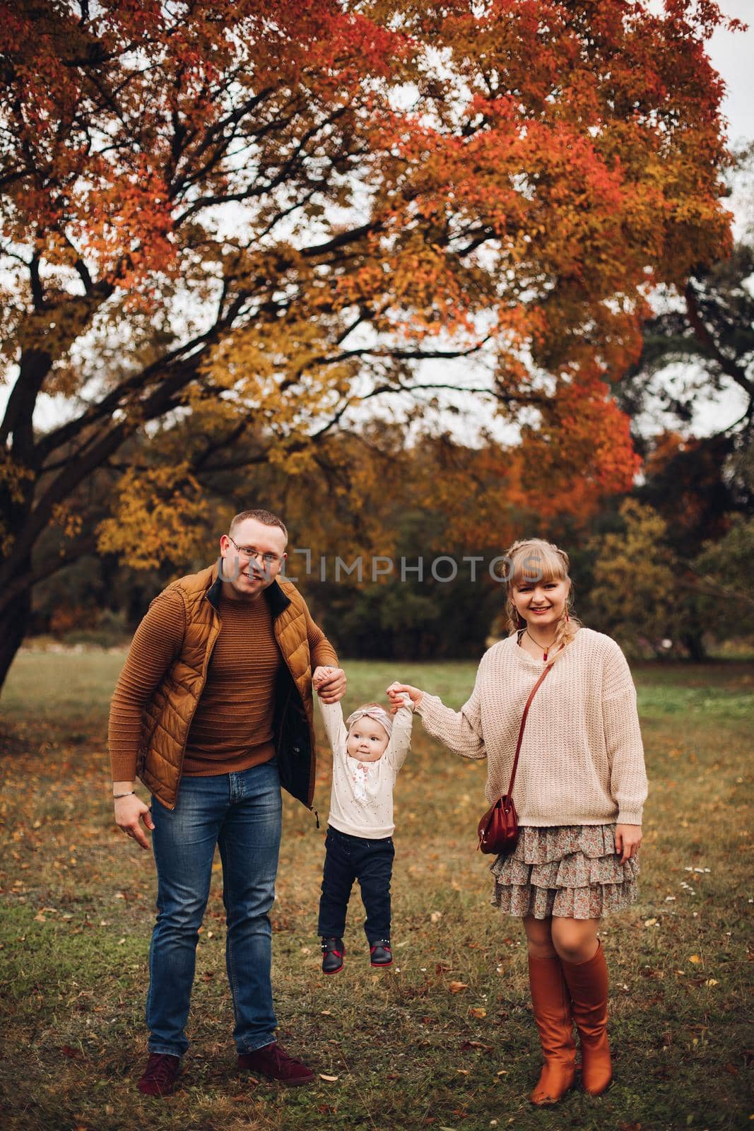 Portrait of attractive young mother and handsome smiling father wearing glasses holding their beautiful lovely baby girl on hands standing against green hedge in autumnal park. They are smiling and looking at camera.