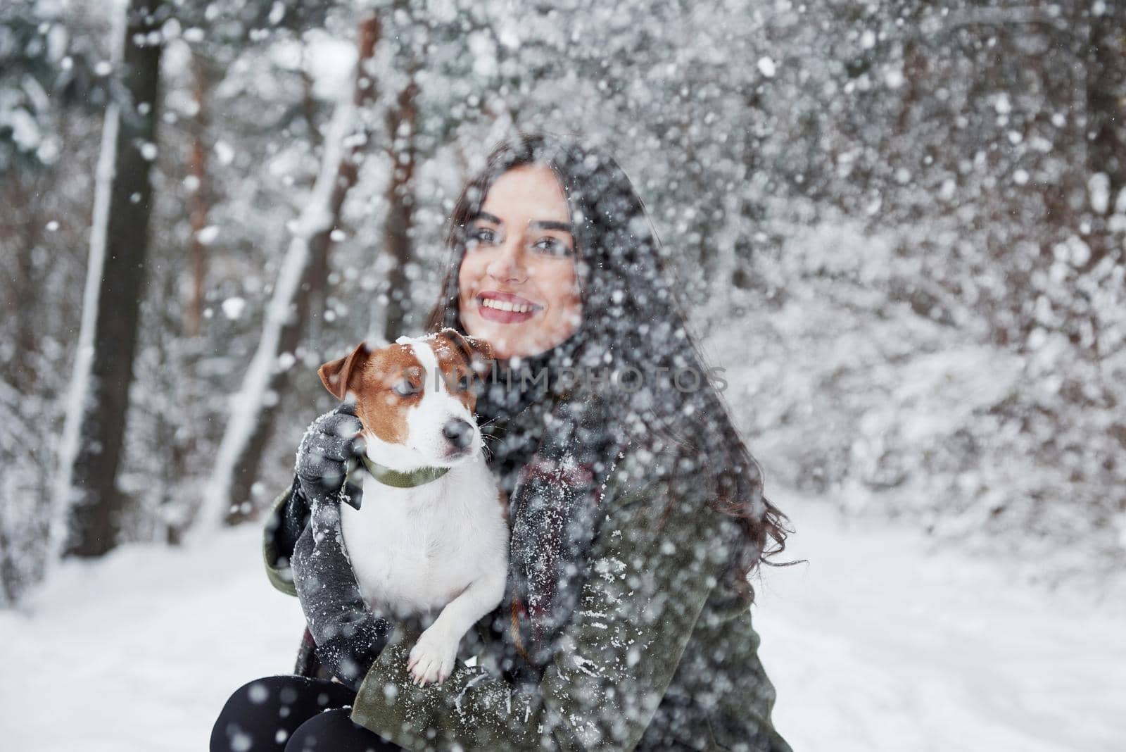 Falling snow. Smiling brunette having fun while walking with her dog in the winter park.