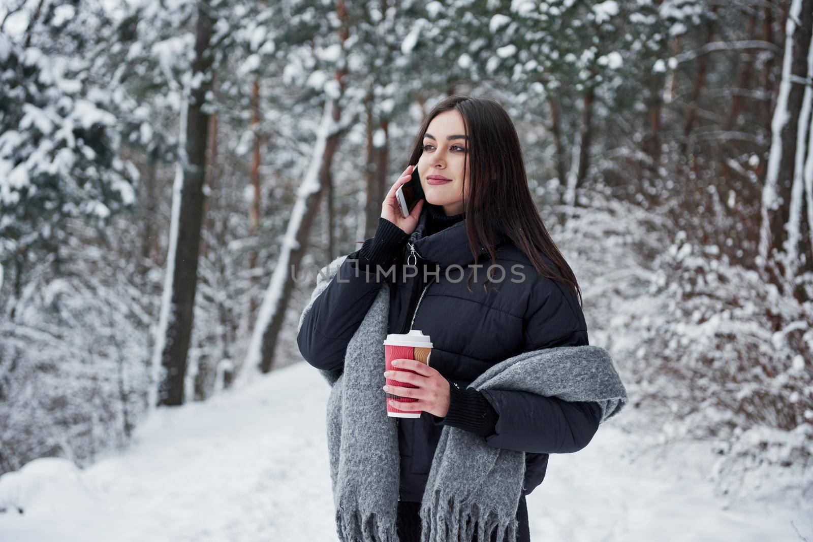 Having conversation on the phone. Girl in warm clothes with cup of coffee have a walk in the winter forest.