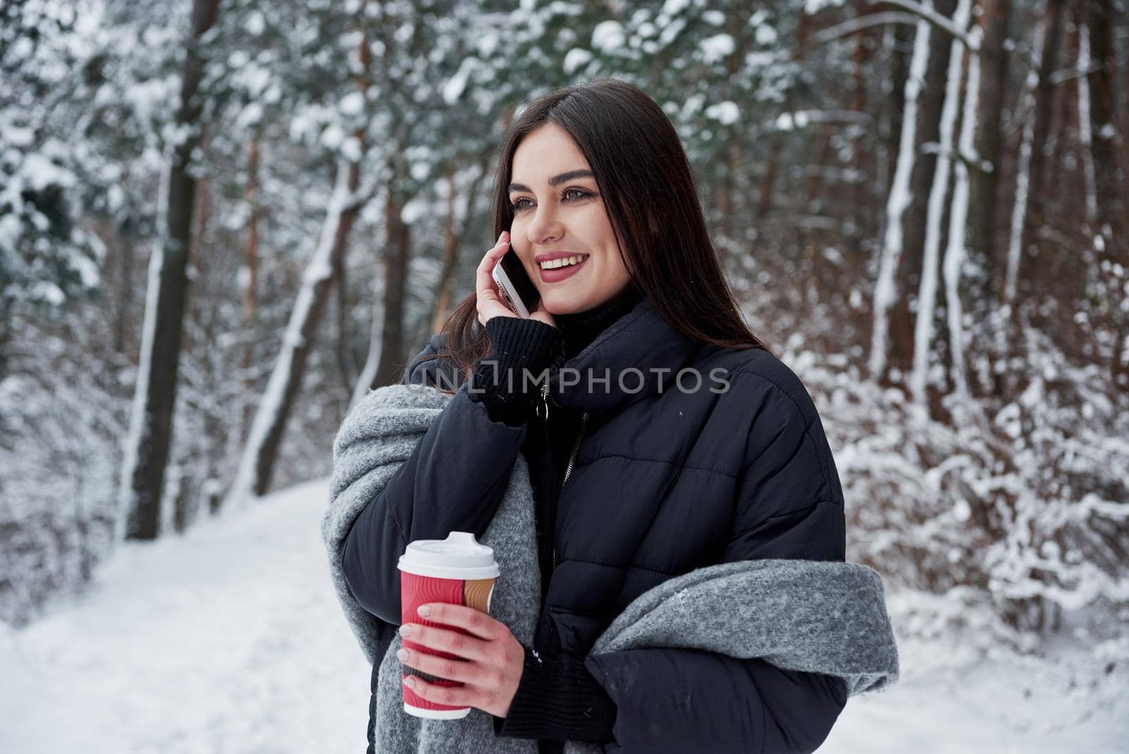 Calling to the friend. Girl in warm clothes with cup of coffee have a walk in the winter forest.