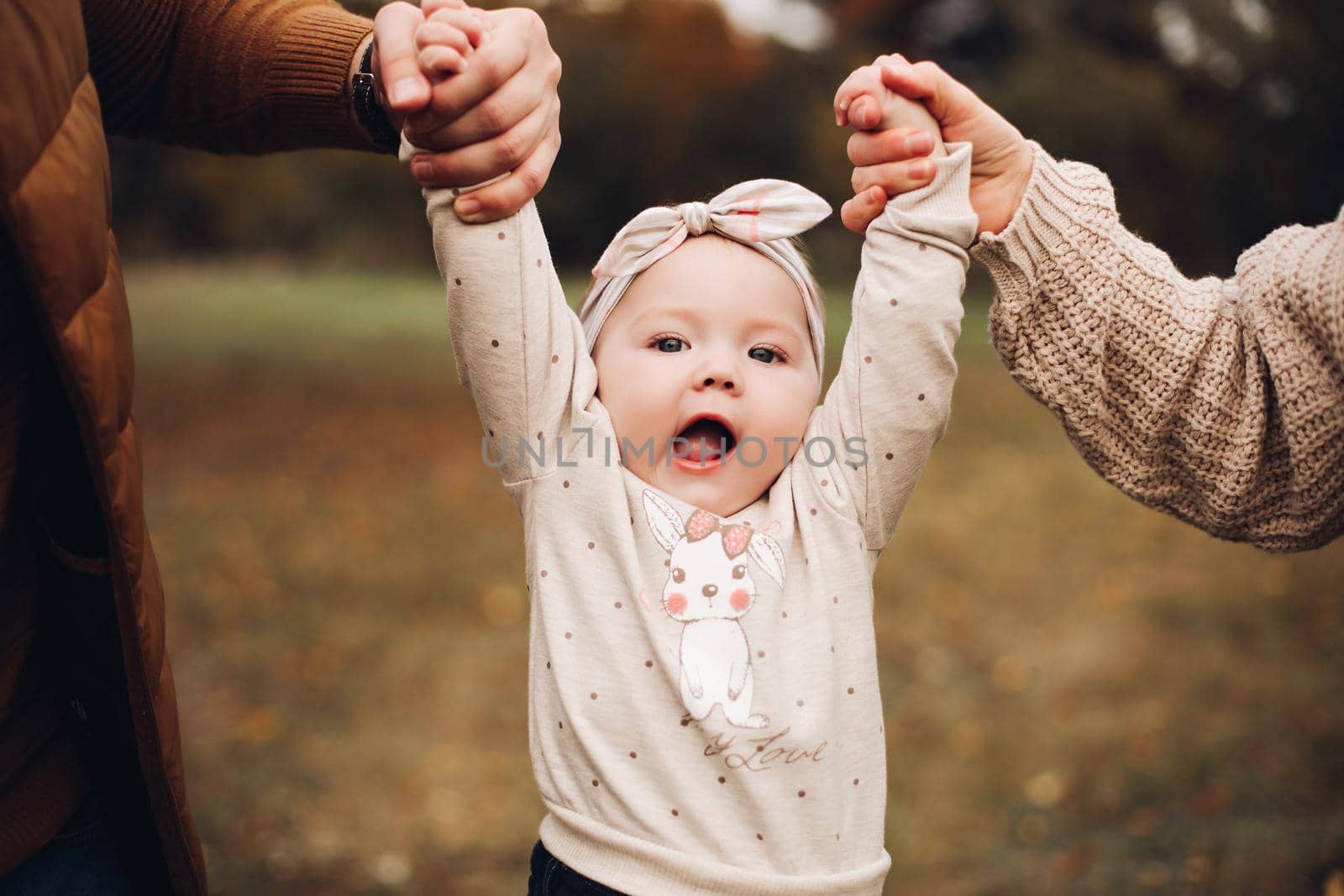 Portrait of lovely little baby girl wearing cute jersey with bunny on it and beige patterned bow headband. She is smiling at camera while parents holding her arms. Weekend in the park in autumn. Baby doing her first steps.
