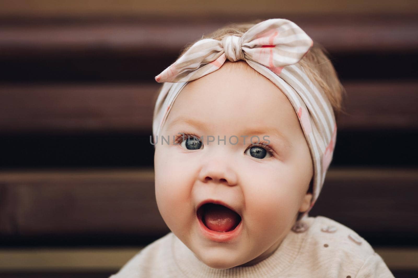Portrait of lovely little baby girl wearing cute jersey with bunny on it and beige patterned bow headband. She is smiling at camera while parents holding her arms. Weekend in the park in autumn. Baby doing her first steps.