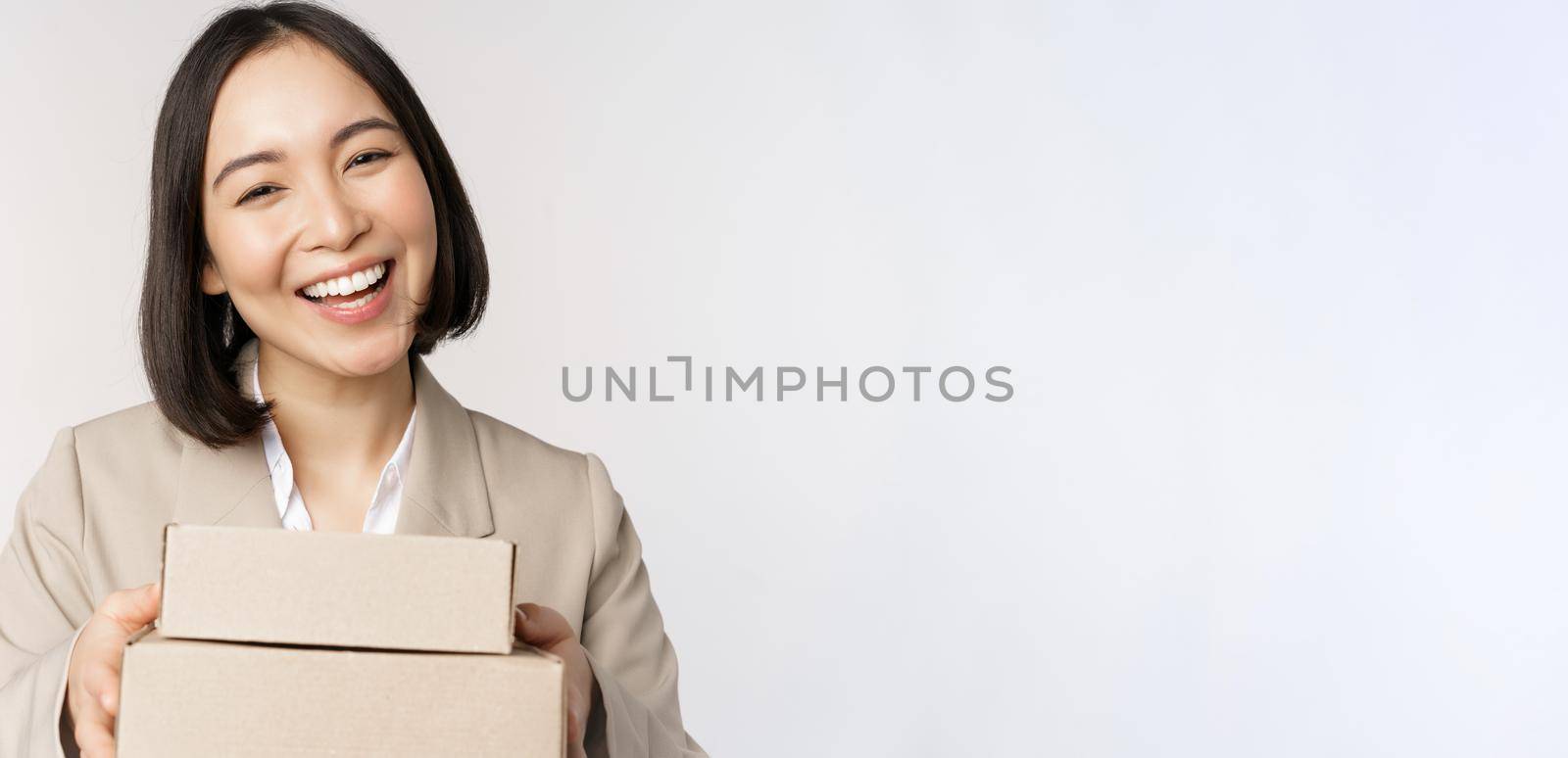 Image of asian saleswoman, business woman giving boxes with order, deliver to customer, standing in suit over white background.