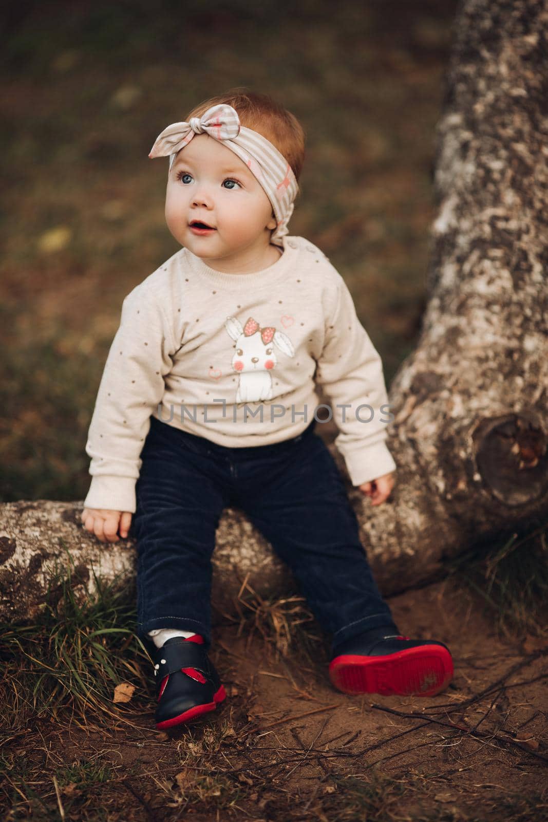 Portrait of lovely little baby girl wearing cute jersey with bunny on it and beige patterned bow headband. She is smiling at camera while parents holding her arms. Weekend in the park in autumn. Baby doing her first steps.